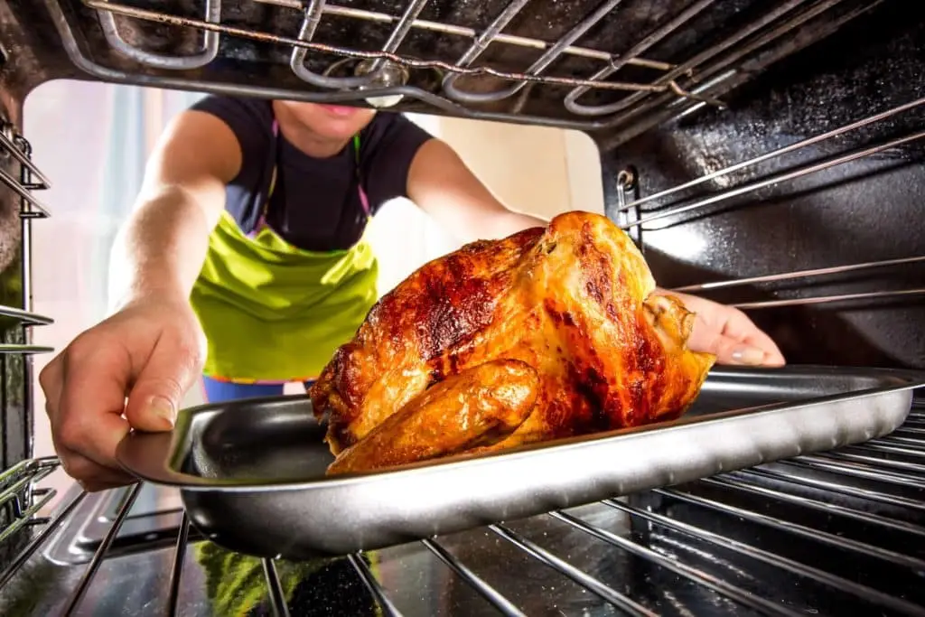 A home cook in an apron carefully slides a golden-brown roasted chicken out of the oven, indicating a moment of delicious triumph in the kitchen.