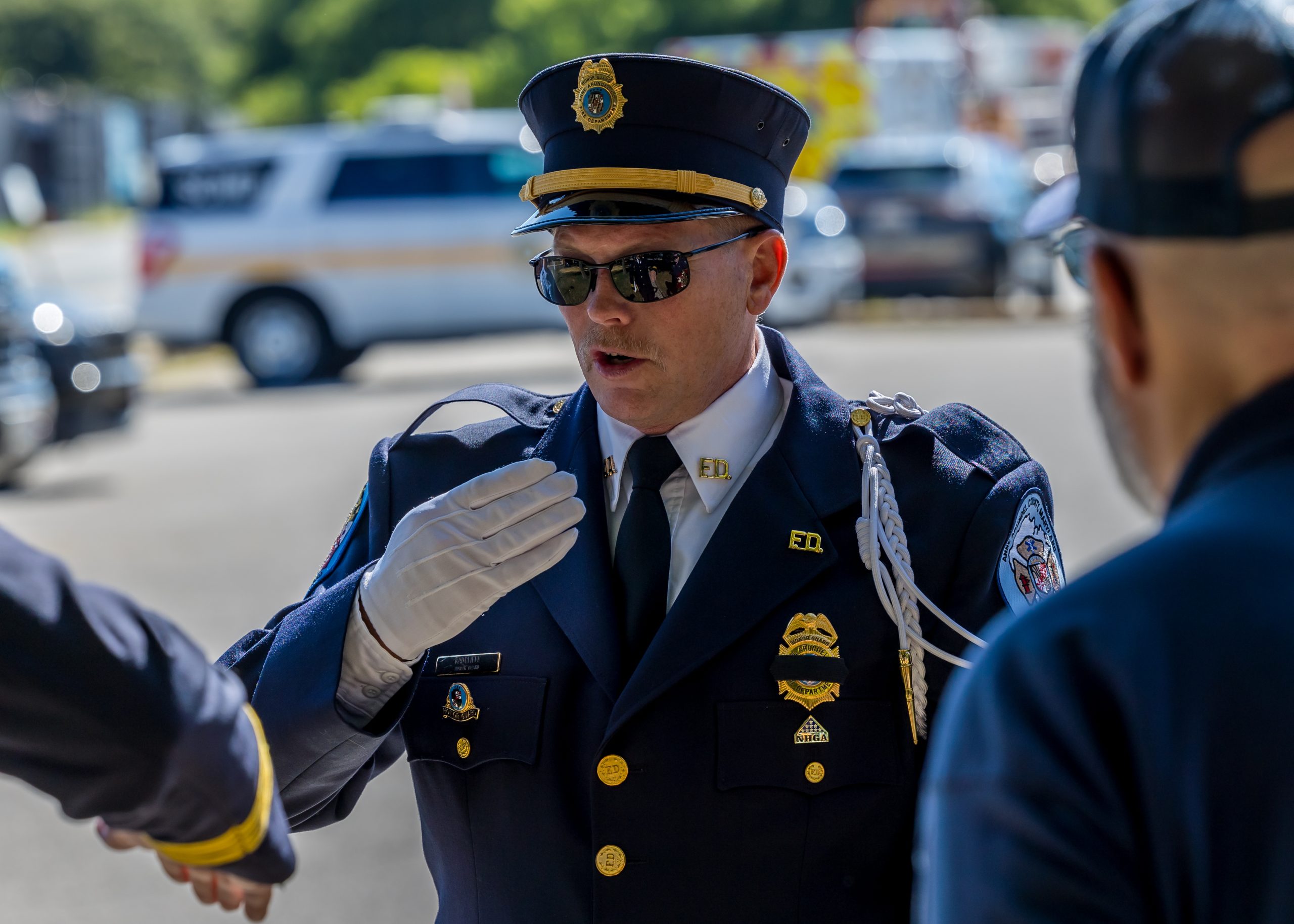 A police officer in formal uniform, including a cap with a badge, dark sunglasses, and white gloves, gestures while speaking. Two other people in similar attire are partially visible. Police vehicles are parked in the background.
