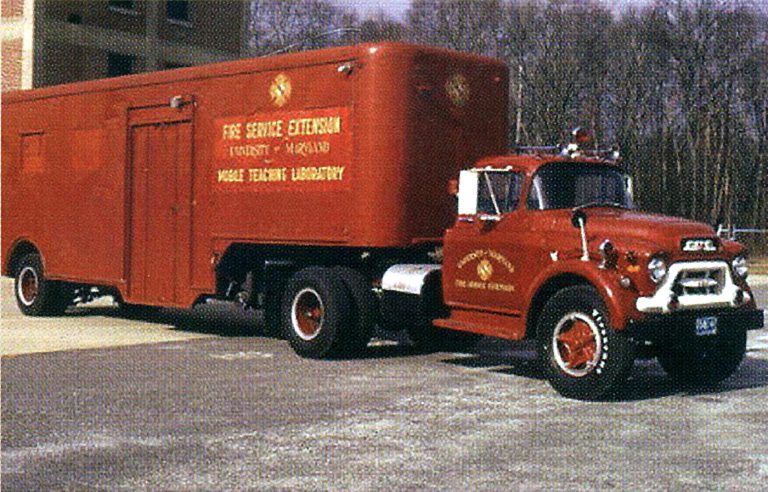 A vintage red fire service truck with a large trailer labeled "Fire Service Extension, University of Maryland, Mobile Training Laboratory" is parked on a concrete surface. Trees and a building are visible in the background.