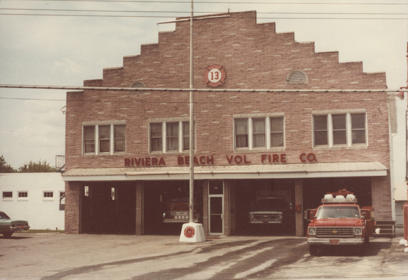 A vintage fire station building labeled "Riviera Beach Vol Fire Co" with three open garage bays. Fire trucks and an emergency vehicle are visible inside. The building has a brick facade with a "13" emblem above.