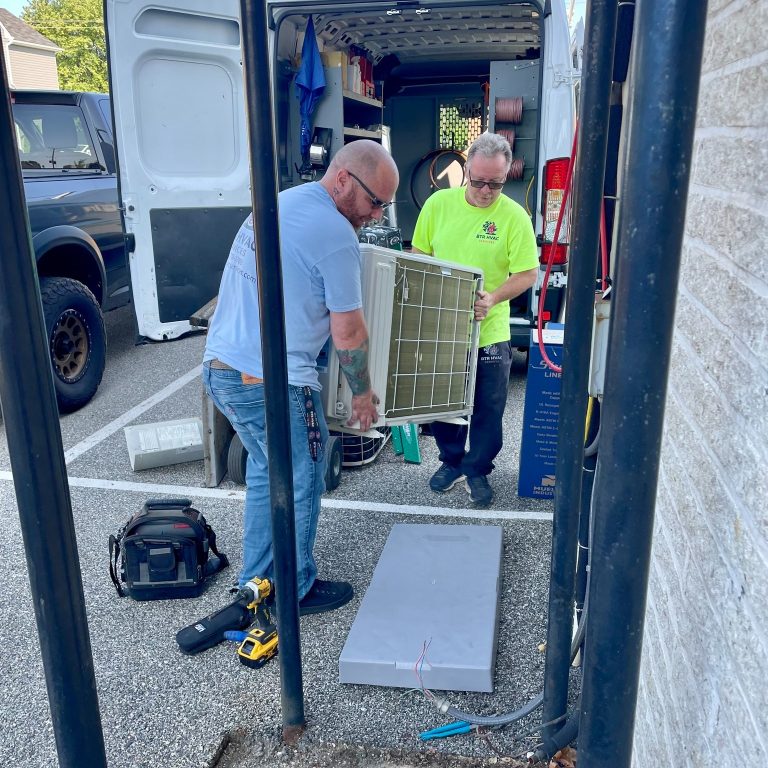 Two men are installing or repairing an air conditioning unit outside. They are standing near an open van filled with tools and equipment. One man wears a light blue shirt, and the other wears a bright yellow shirt. Tools are scattered on the ground.