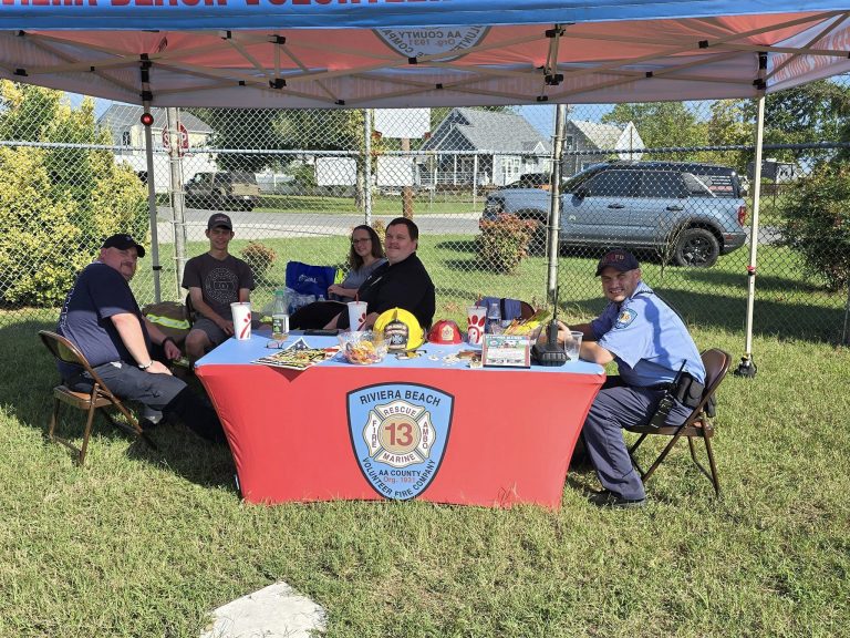 A group of five people sitting under a tent with a red tablecloth displaying the Riviera Beach Volunteer Fire Company logo. The table holds various items, and a street with houses and cars is visible in the background.