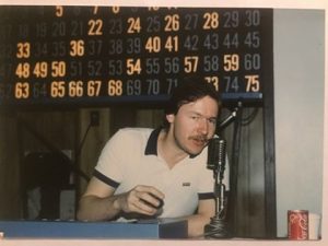 A man with a mustache sits at a table with a microphone, in front of a bingo board displaying numbers. A can of Coca-Cola is on the table. He appears to be speaking or announcing.