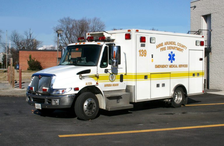 A white and yellow ambulance from Anne Arundel County Fire Department is parked outside a building. The vehicle has the emergency medical services emblem and number 139 on its side. It's a clear day with a few trees and a brick building in the background.