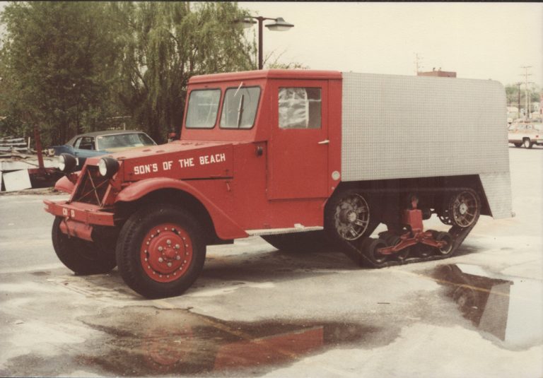 A vintage red truck with "Son's of the Beach" painted on the side. The vehicle has an unusual rear, featuring large treads instead of traditional wheels, parked on wet pavement with trees and a parked car in the background.