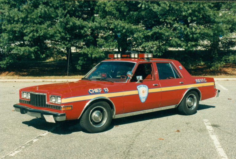 A vintage red fire chief car with "CHIEF 13" written on the side, parked in an empty lot. The car has a light bar on top and a shield emblem on the door. Pine trees are visible in the background.