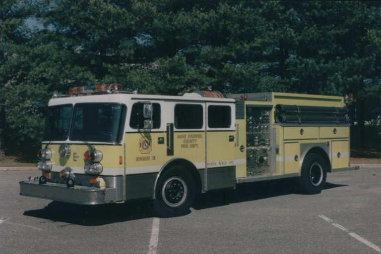 A yellow and white fire truck parked in an empty lot, surrounded by trees. It is labeled "Anne Arundel County Fire Dept." and features various lights and equipment compartments.