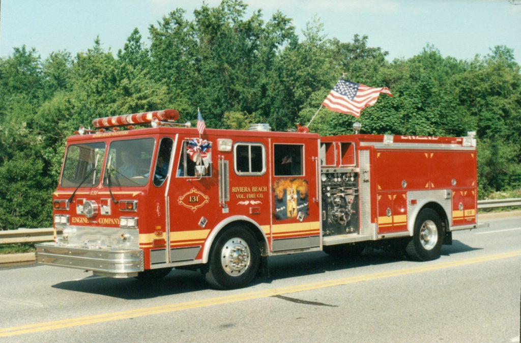 A red fire truck with "Engine Company 131" and "Riviera Beach Volunteer Fire Co." on the side is driving on a road. It has American flags attached to it, with trees in the background.