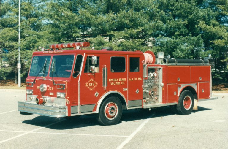 A red fire truck is parked in an empty lot with trees in the background. The truck features equipment compartments and hoses, with a text on the side indicating "Riviera Beach Vol. Fire Co.