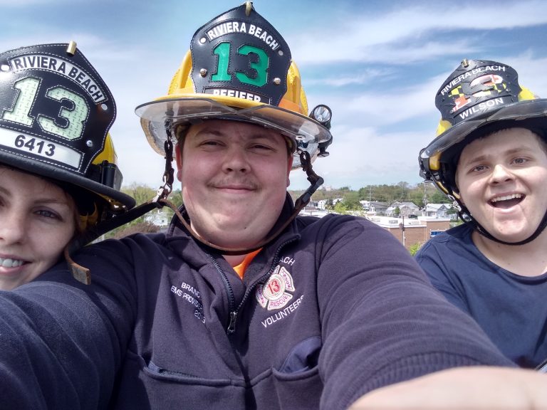 Three firefighters in uniform and helmets smile at the camera, posing for a selfie outdoors. The background features a clear sky and distant buildings.