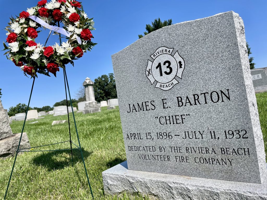 A gravesite features a gray headstone for James E. Barton, labeled "Chief," dated April 15, 1896 - July 11, 1932. It's dedicated by the Riviera Beach Volunteer Fire Company. A floral wreath with red and white flowers stands nearby.