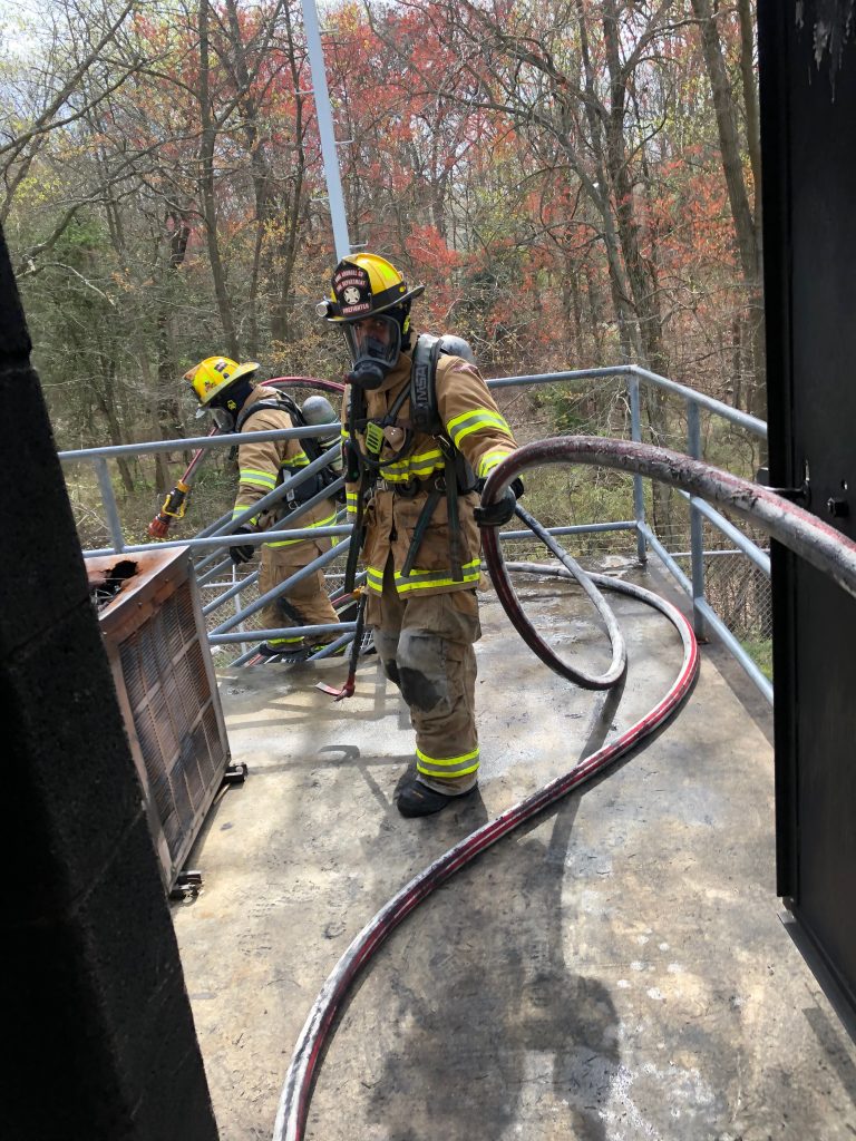 Two firefighters in full gear, including helmets and oxygen masks, handle a fire hose on a metal platform surrounded by trees with autumn foliage. One firefighter holds onto the hose while the other is adjusting equipment.