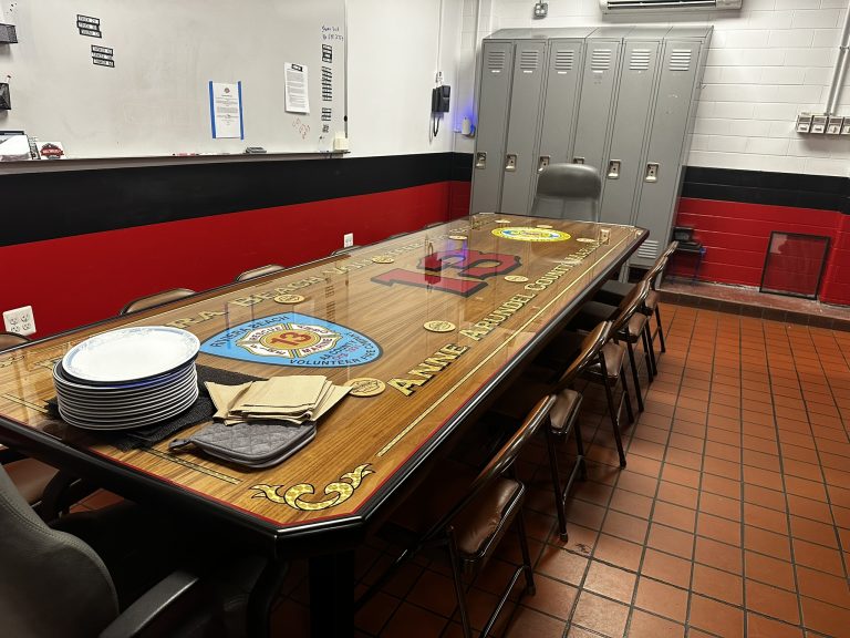 A meeting room with a large table featuring logos and the words "Anne Arundel County." Plates and napkins are on one end. Surrounding chairs are placed on both sides. Lockers line the back wall, and the room has red and black walls with white accents.