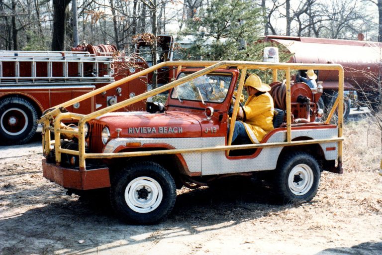 A vintage red fire service jeep labeled "Riviera Beach" is parked outdoors. It features yellow safety bars and firefighting equipment. A person in a yellow uniform is seated in the vehicle. Trees and another fire truck are in the background.