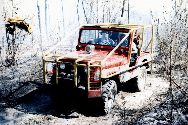 A red off-road vehicle with a protective yellow roll cage navigates through a sparse, wooded area, stirring up dust. The driver is focused on the path ahead, surrounded by bare trees and dry underbrush.
