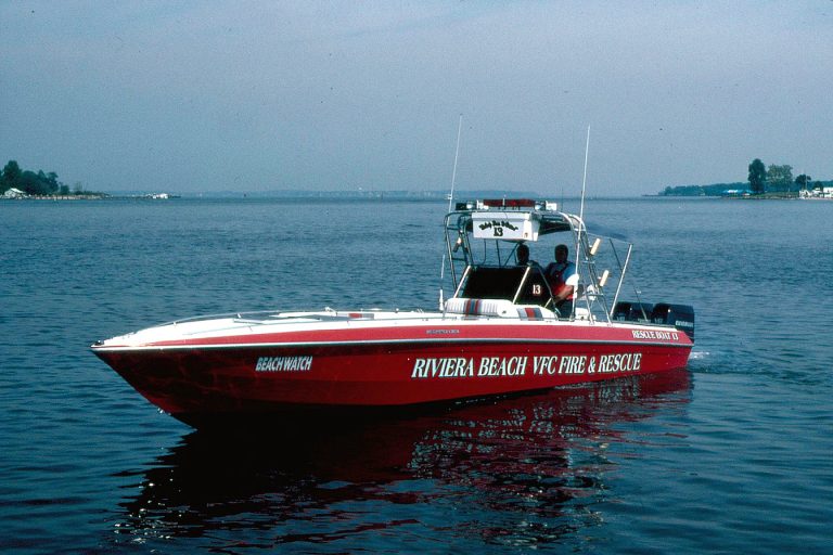 A red fire and rescue boat with "Riviera Beach VFC Fire & Rescue" written on the side is on calm water. The boat has antennas and is manned by a person in uniform. There's a distant shoreline with trees in the background.
