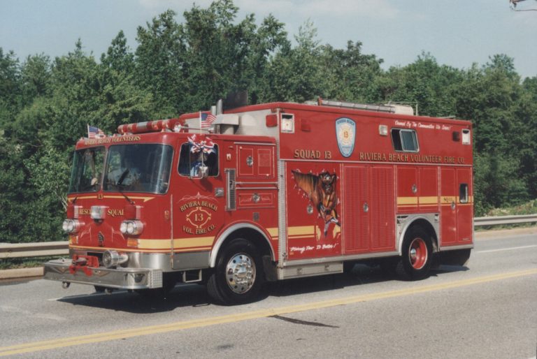 A red fire truck with "Squad 13" and "Riviera Beach Volunteer Fire Co." on its sides is parked on a road. The truck is decorated with American flags and features a detailed emblem and mural. Trees are visible in the background.