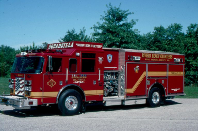 A red fire truck with "Squad 11A" and "Rivera Beach Volunteers" written on the side, parked on a paved surface. Trees and a clear blue sky are in the background.