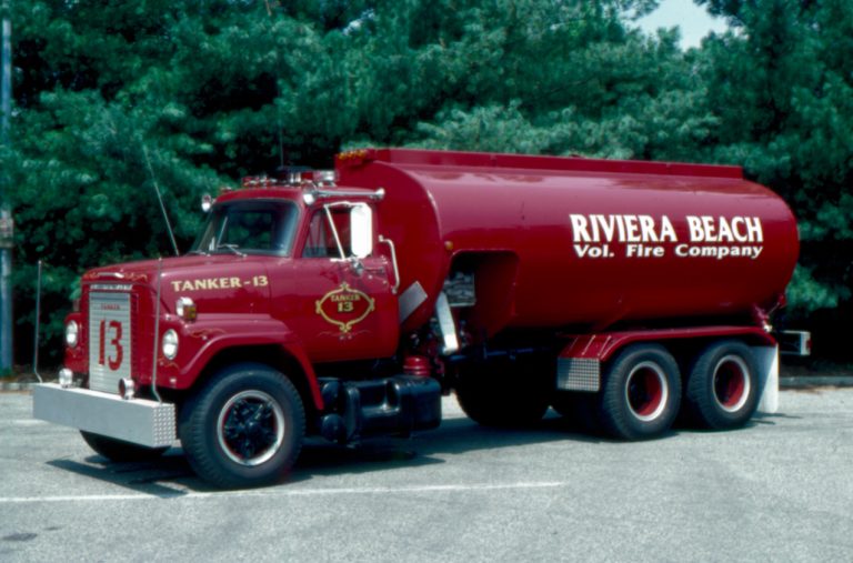 A vintage red fire tanker truck with "Riviera Beach Vol. Fire Company" and "Tanker 13" written on it, parked on a road with green trees in the background.