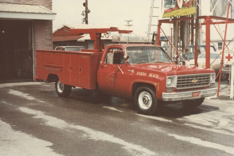 A red utility truck with "Riviera Beach" and "Fire Rescue Unit 13" on the side is parked outside a building. The truck has a cherry picker on its bed, and a ladder is visible in the background.