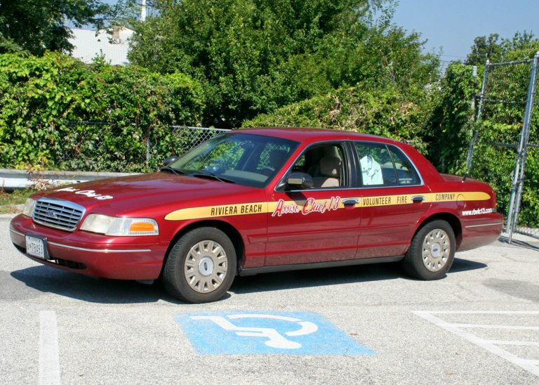 A red volunteer fire department car, labeled "Riviera Beach" and "Anne Arundel County 13," is parked in a handicapped space. The car has yellow and black stripes along its sides. Trees and a fence are visible in the background.