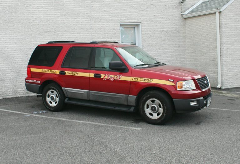 A red SUV marked with "Chief" on the door and "Volunteer Fire Company" on a yellow stripe is parked in an empty lot next to a white brick wall. The vehicle has a roof rack and website address on the rear side window.