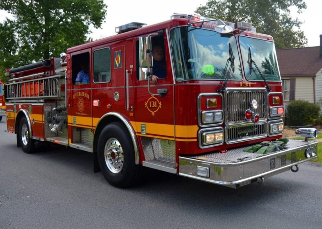 Red fire truck with yellow stripes, marked "131" on the side, parked on a residential street. A person is visible inside the driver's seat. Nearby are green hoses and a house with trees in the background.
