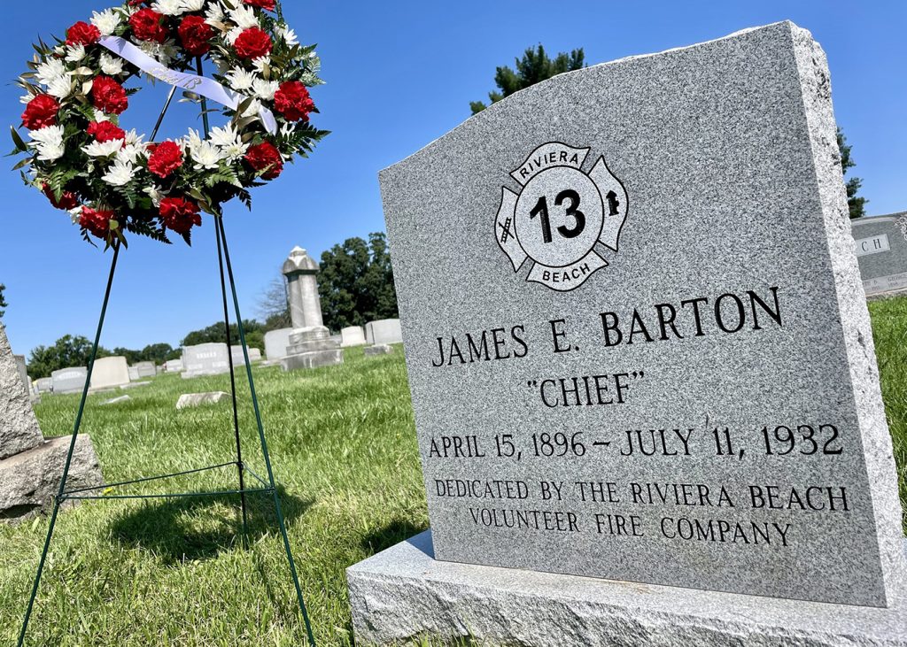 A gravestone in a cemetery with a firefighter emblem engraved on it reads "James E. Barton, 'Chief,' April 15, 1896 - July 11, 1932." Nearby, a wreath of red and white flowers stands on a frame. Lush green grass and other gravestones surround the area.