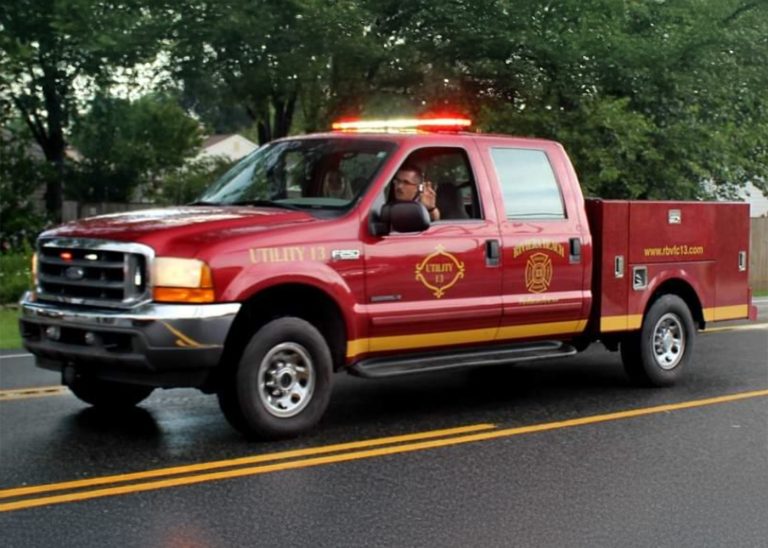 A red utility fire department truck with "Utility 13" marked on the side is driving on a road. The vehicle has emergency lights on, and there is a person in the driver's seat. Trees and houses are visible in the background.