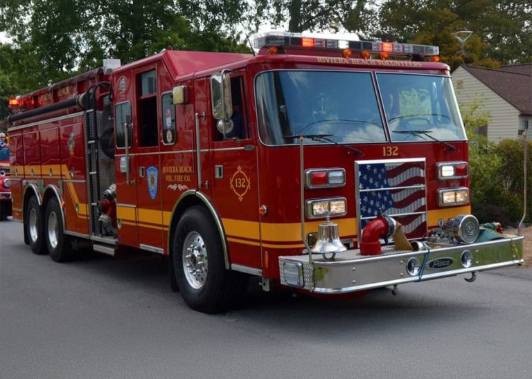 A red fire engine with "132" labeled on its side is parked on a street. It features an American flag design on the front grille, with trees and a residential area in the background.
