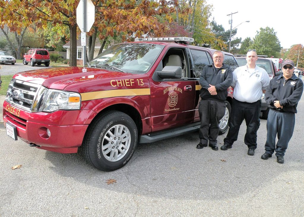 Three people stand beside a red fire department SUV labeled "Chief 13" on a tree-lined street. The group includes two men in uniform and one in a jacket. The background shows autumn foliage and parked cars.
