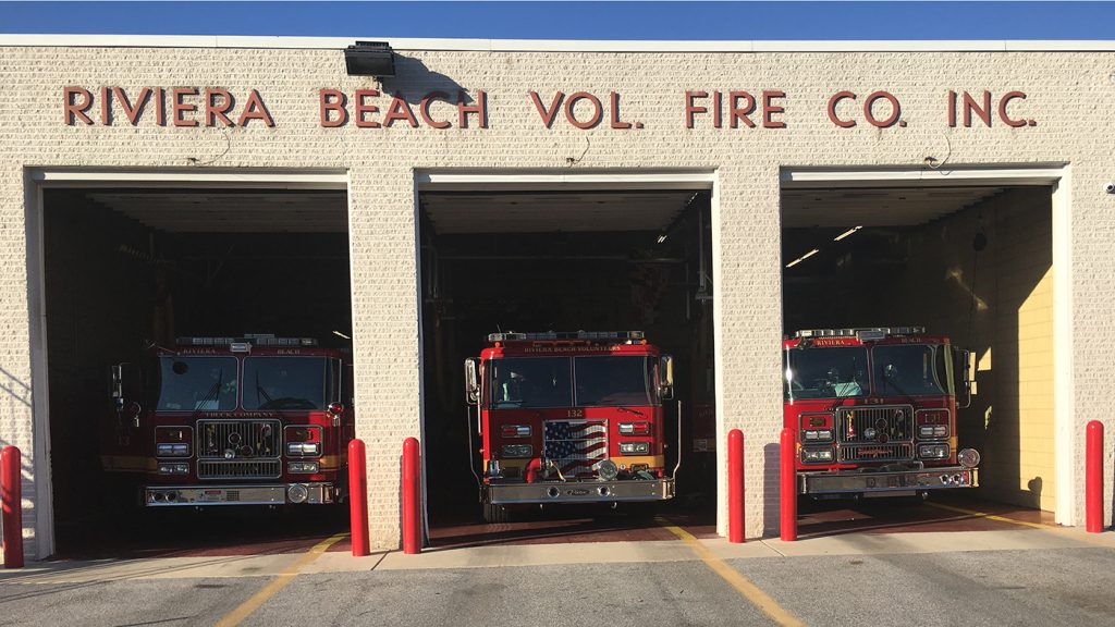 Three red fire trucks are parked in the bays of the Riviera Beach Volunteer Fire Company building. The firehouse is made of light-colored brick, and the company name is displayed in red lettering above the bays.