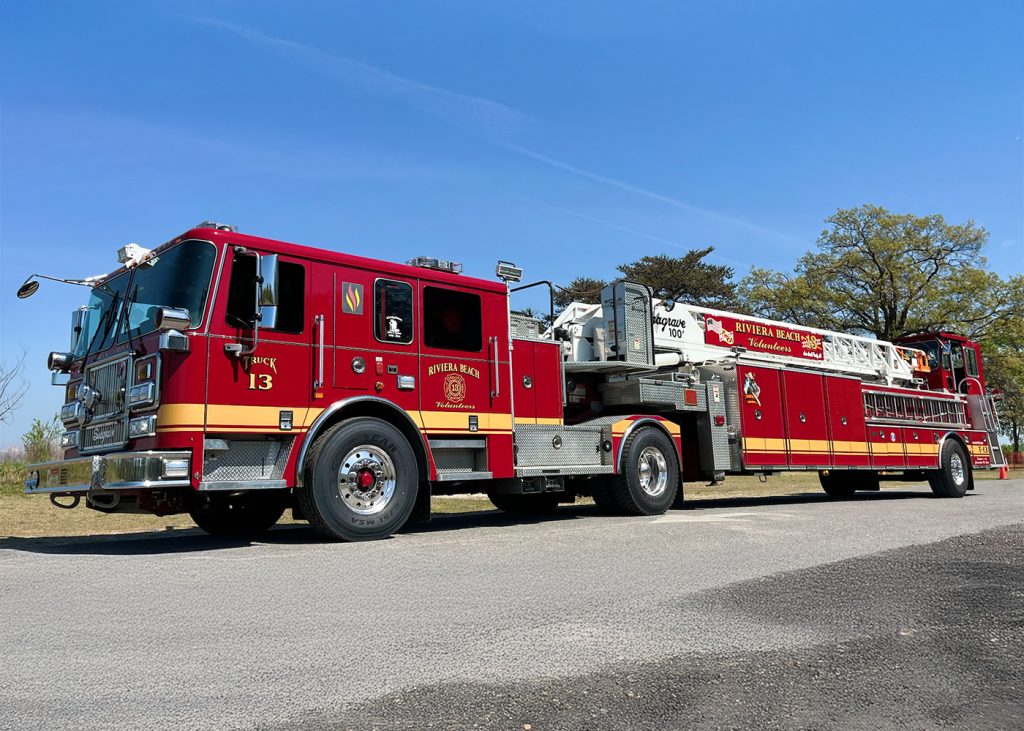 A red and yellow fire truck labeled "River Rescue" with a ladder extended sits on a paved road under a clear blue sky, surrounded by green trees. The truck is marked with the number 13 and has various equipment compartments.