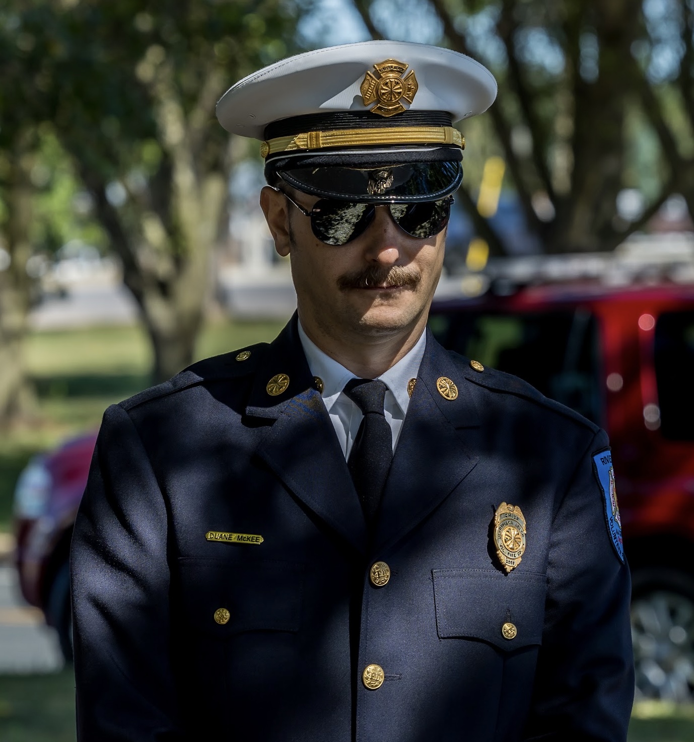 A person in a navy blue uniform with a badge and a white hat stands outdoors. They wear sunglasses and a mustache. Trees and a red vehicle are in the blurred background.