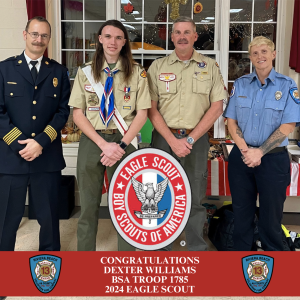 A group photo of four people standing indoors. The person on the left is in a fire department uniform, two in the middle are in scout uniforms, and the person on the right is in a different uniform. A "Boy Scouts of America" emblem is visible.