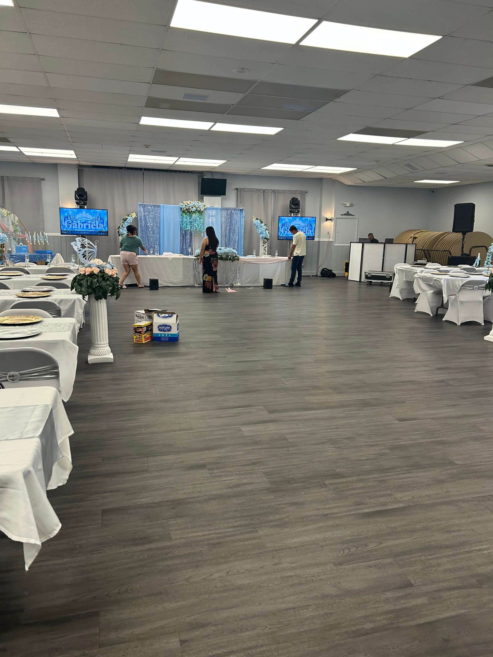 A large event hall with gray flooring, white-clothed tables along the sides, and three people setting up decorations in the center. Floral arrangements, TV screens, and banquet chairs are visible around the room.