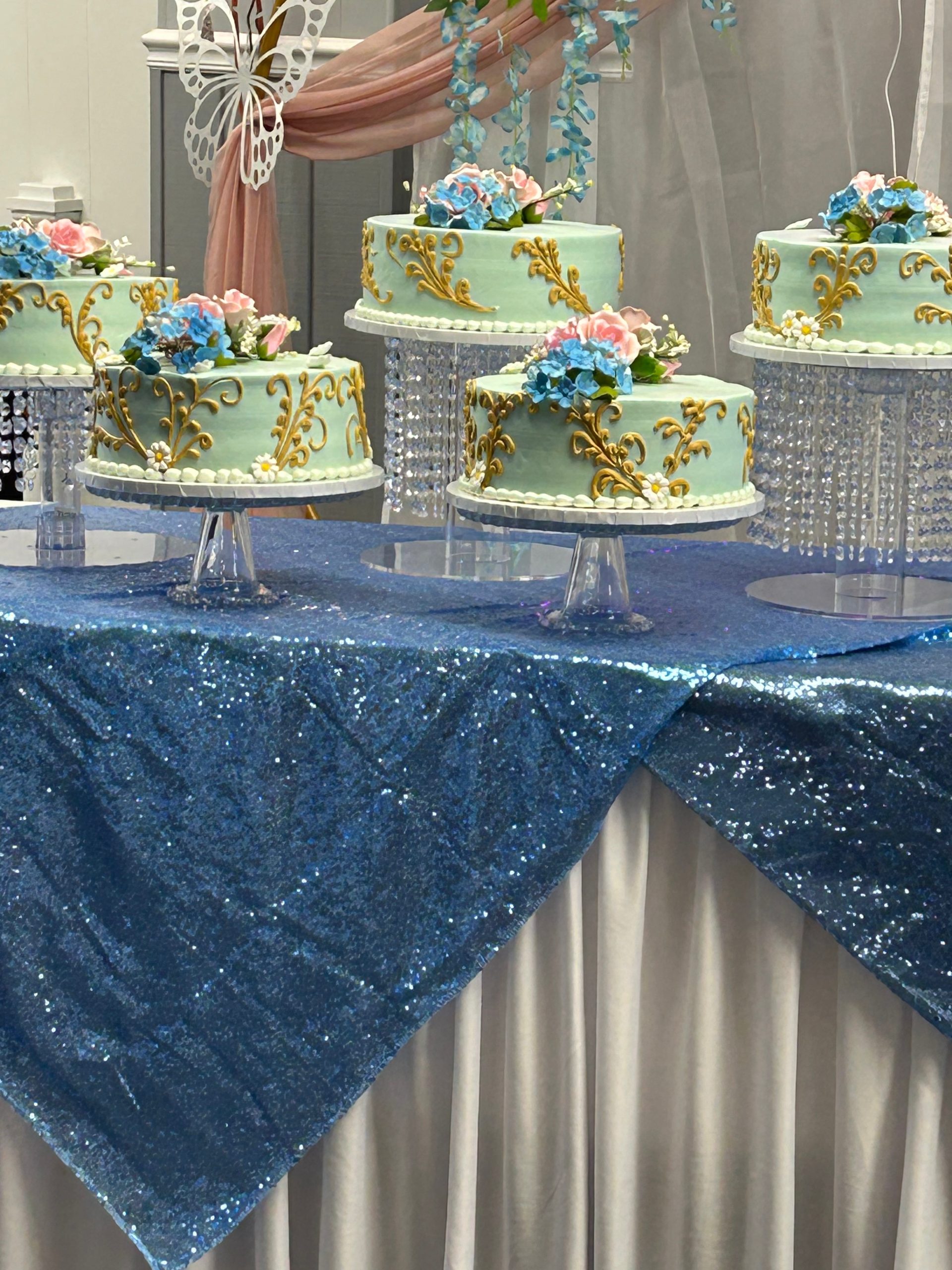 Several ornate cakes with gold decorations and floral toppers are displayed on crystal stands atop a shimmering blue tablecloth at a decorated event. A butterfly ornament and pink drapery are in the background.